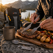 Load image into Gallery viewer, A person outdoors at sunset is cutting a grilled steak on a wooden board with camping utensils, with an APEX backpack, metal mug, spoon, and knife visible in a mountain-and-lake setting.