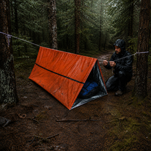 Load image into Gallery viewer, A hiker in a black raincoat kneels beside an orange APEX survival shelter setting it up between trees in a forest.
